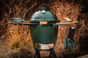 Ein Big Green Egg Medium mit Holzablagen, Grillhandschuh, Zange und einem frisch gegrillten Sandwich in herbstlicher Umgebung.