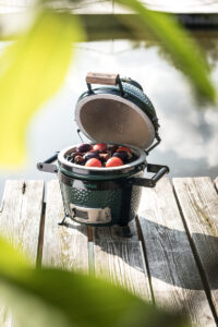 Ein Big Green Egg Mini steht geöffnet auf einem Holzsteg am Wasser und ist mit frischen Auberginen und Tomaten gefüllt.