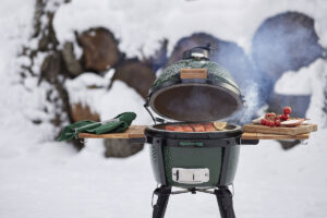 Ein Big Green Egg MiniMax steht im Schnee und grillt ein saftiges Lachsfilet mit Zitrone, während Rauch aufsteigt.