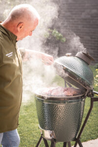 Ein Grillmeister in grüner Kochjacke öffnet den rauchenden Big Green Egg Kamado-Grill, während auf dem Rost saftiges Fleisch gegart wird.