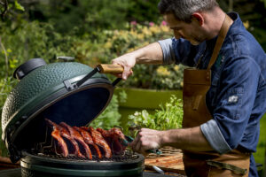 Ein Grillmeister mit Lederschürze öffnet den Big Green Egg Kamado-Grill und überprüft die langsam gegarten Spareribs.